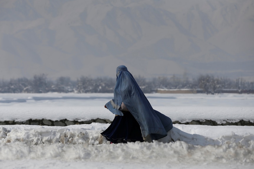 An Afghan woman wearing a burqa walks along a street covered with snow.