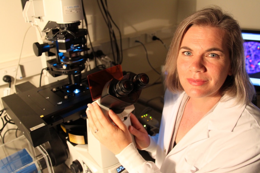 A woman with blonde hair and a white jacket sitting in front of a microscope in a lab.