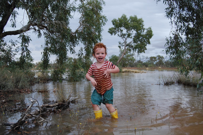 A red haired toddler with a red and white striped shirt and yellow gumboots smiles as she stands in a body of shallow water.