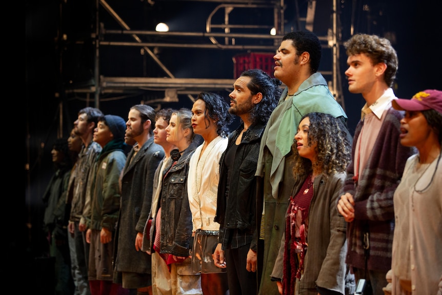 The cast, wearing scrappy clothes, stands in a line singing, the characters of Tom Collins and Angel in the centre