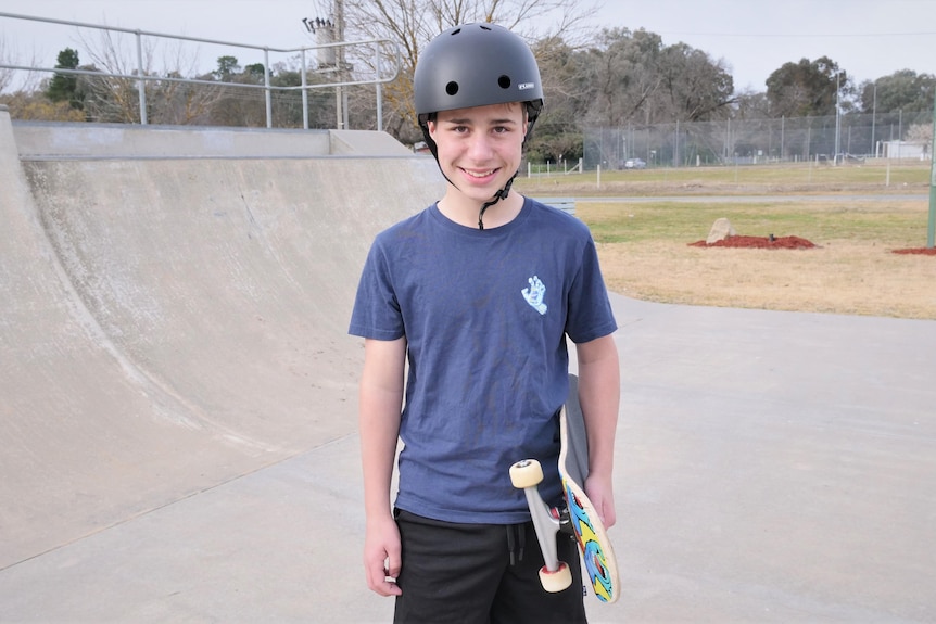 A child standing on a skating half-pipe with his skateboard tucked under his arm 