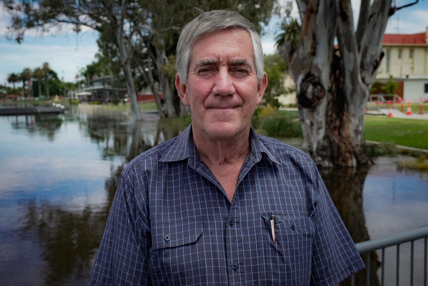 A man looks solemnly at camera with short grey hair, he wears a blue short sleeved button up, behind him is rising flood water