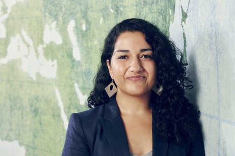 A woman with long dark curly hair, big diamond shaped earring, a navy blazer, stares directly at the camera.