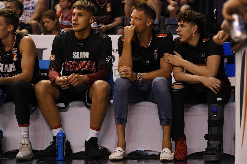 Two NBL players sit on the sidelines, with the player on the right in a moon boot.