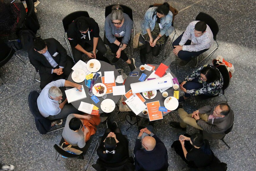 A dozen people sit around a table littered with paperwork, seen from above.