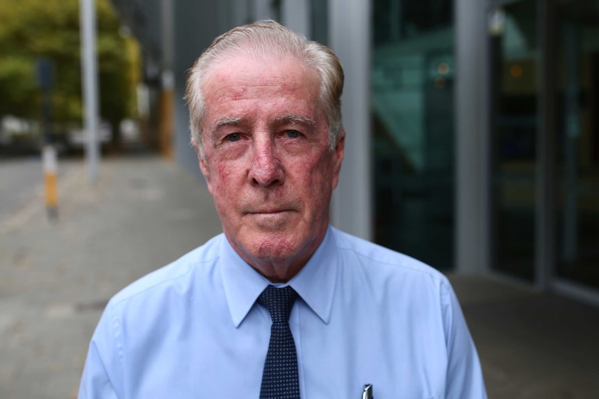 An older man with white hair faces the camera wearing a blue shirt and dark blue tie.