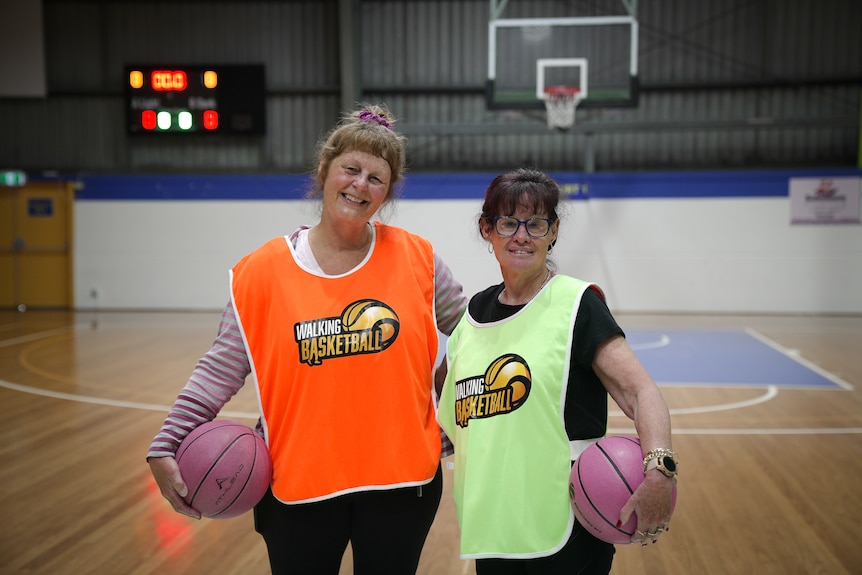 Two women smile while holding pink basketballs and wearing branded walking basketball singlets.