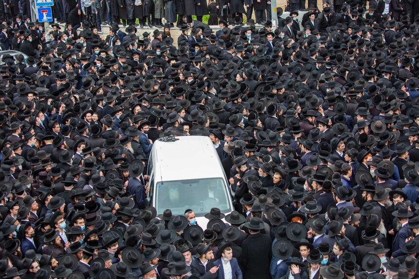 A large group of orthodox Jews with beards and wearing black surround a white car in a city square.