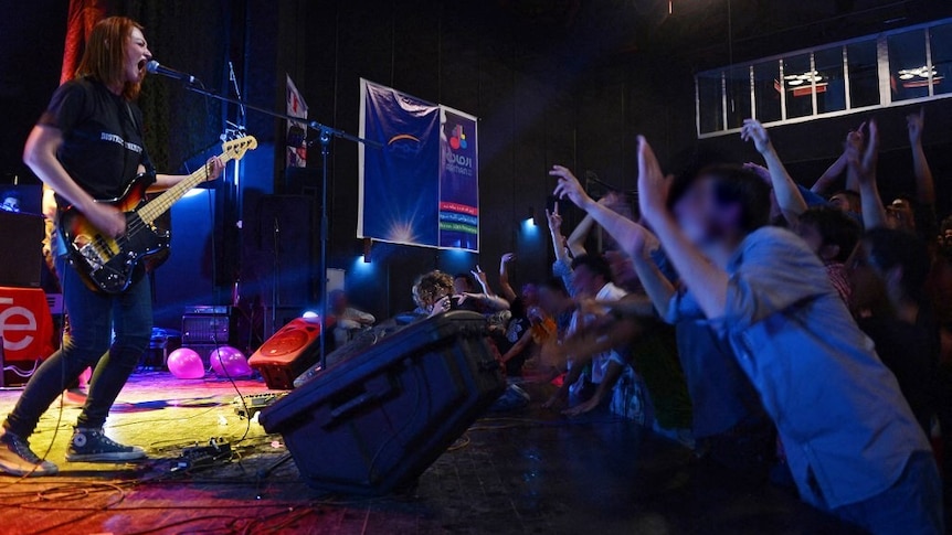 A guitarist sings into a microphone on a stage as crowd members raise their hands