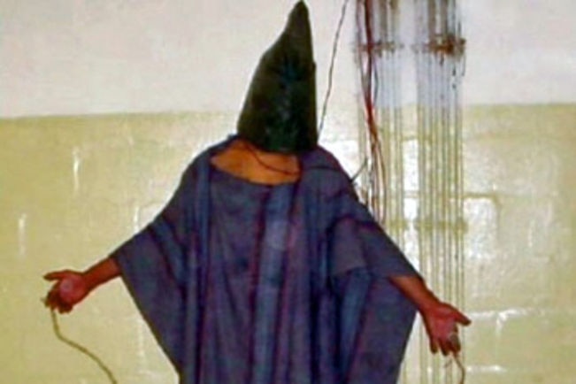 Iraqi detainee standing with a bag on his head, standing on a box with wires attached to his fingers