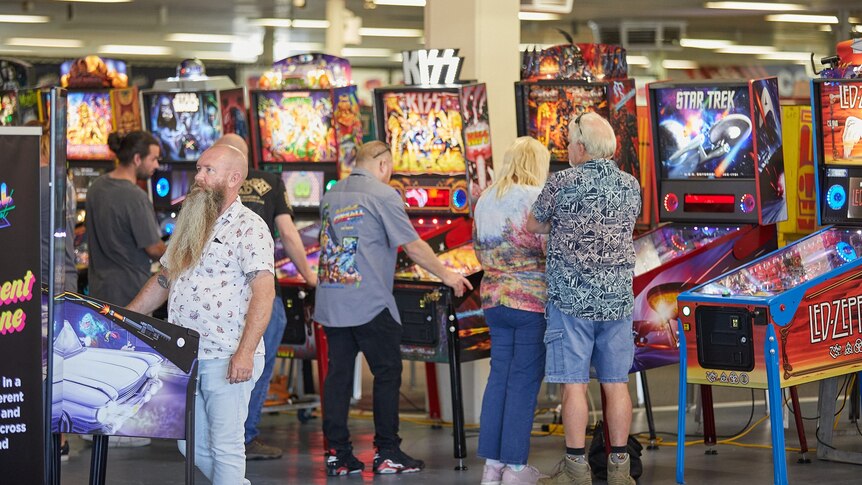 Crowd of people stand around a row of pinball machines, lights flashing