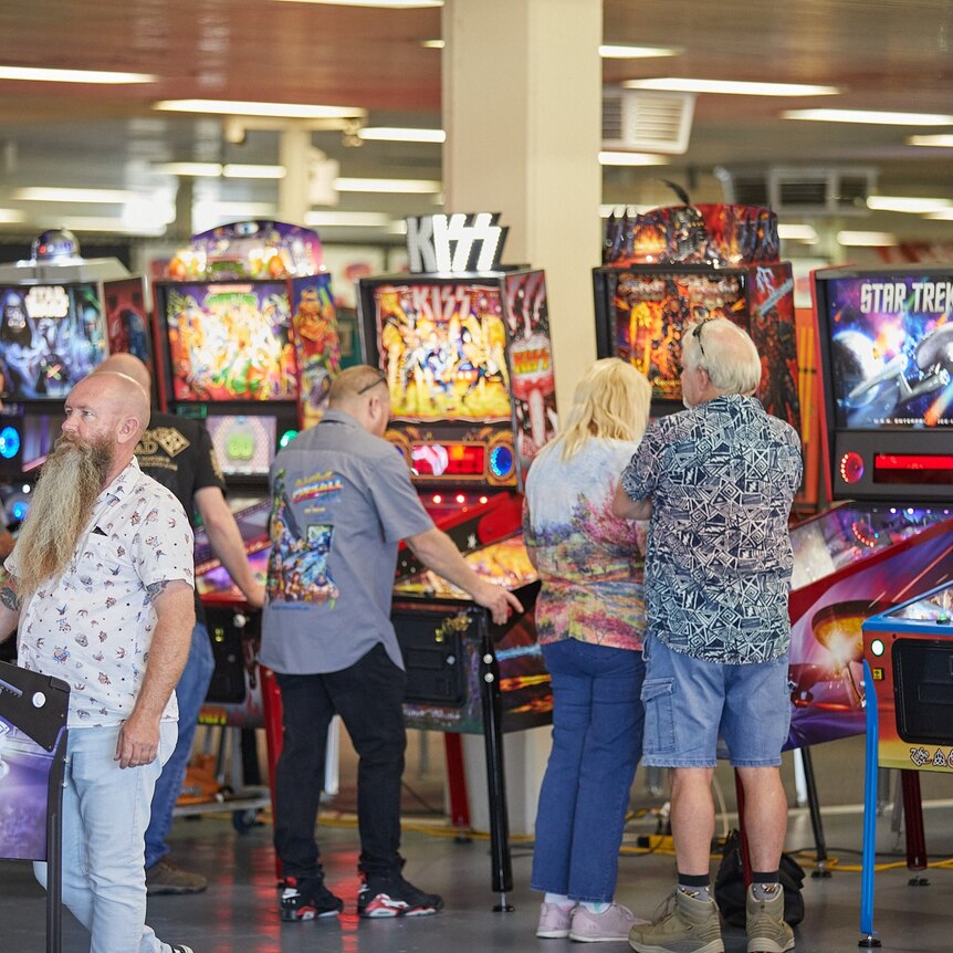 Crowd of people stand around a row of pinball machines, lights flashing