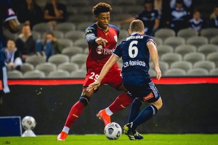 A male soccer player wearing red dribbling a ball next to another man wearing blue