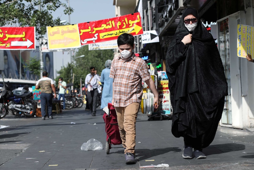 A woman wearing black and a face mask, walks with a mask-wearing boy along shopping district street.