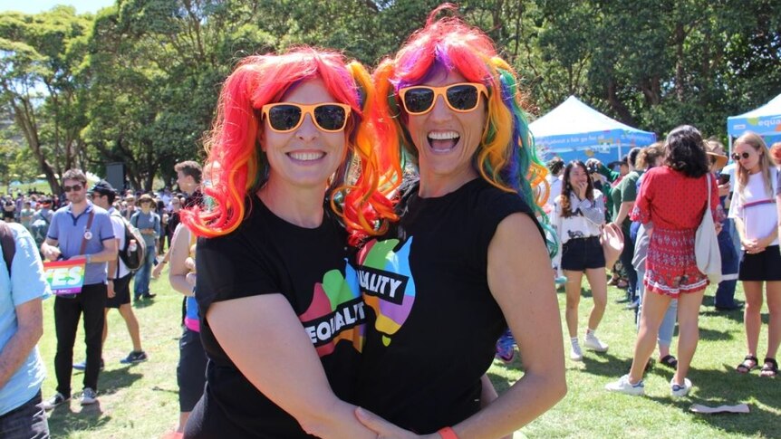 Shalini Schultz and Devon Indig hug wearing rainbow wigs and tutus with orange sunglasses.