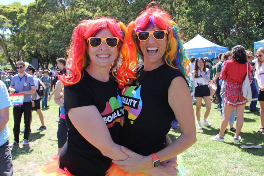 Shalini Schultz and Devon Indig hug wearing rainbow wigs and tutus with orange sunglasses.