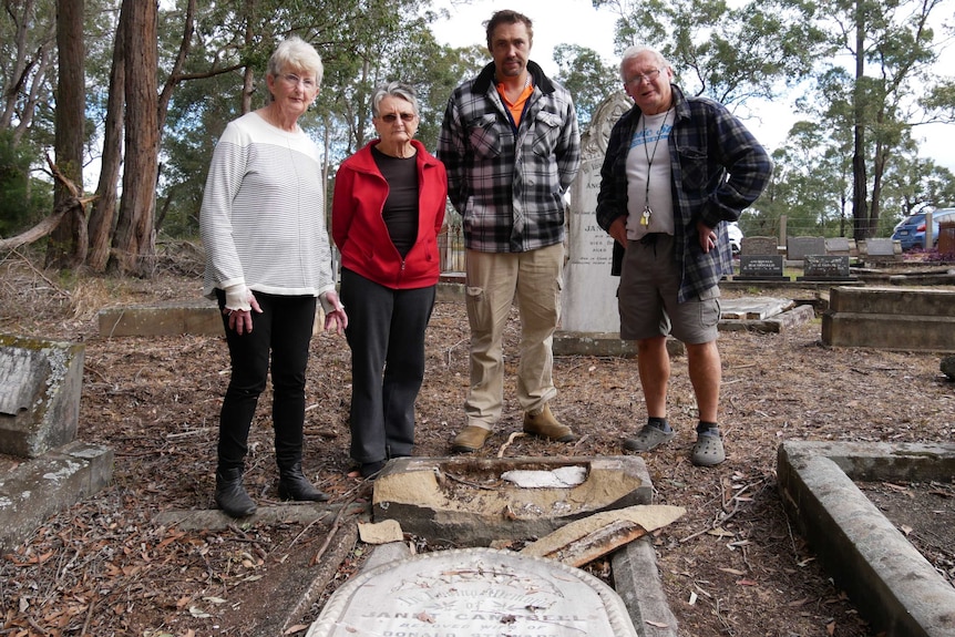 Four people stand around a flattened gravestone looking concerned, at a cemetery surrounded by bushland.
