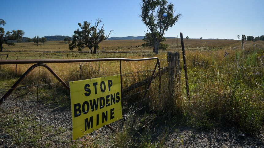 A yellow sign on a metal gate reads 'stop Bowdens mine' in a rural landscape