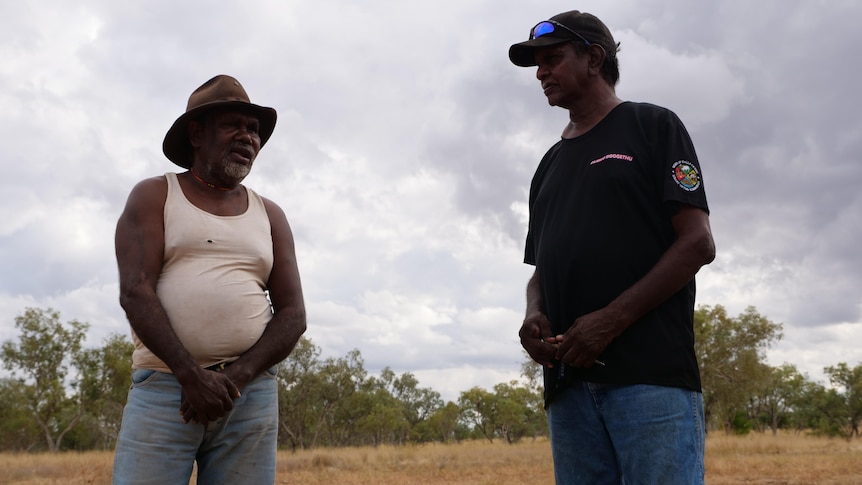 two indigenous men talk in a tree-rimmed bush clearing