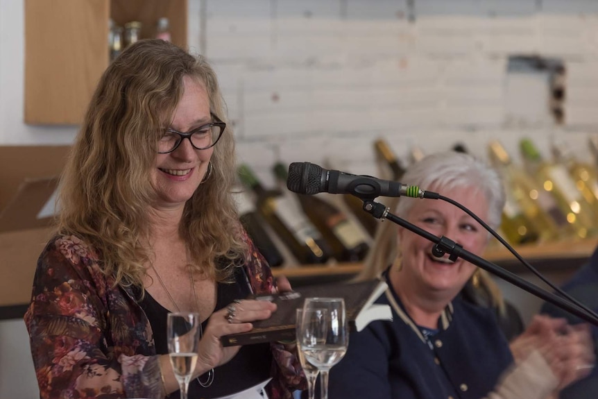 A woman behind a microphone holding a book and smiling as another woman looks on.