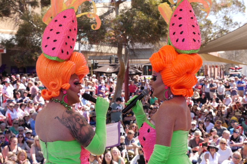 Two drag queens dressed in matching watermelon themed costumes entertaina large crowd outdoors.