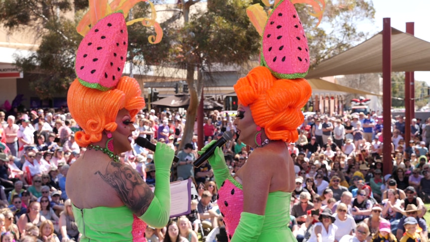 Two drag queens dressed in matching watermelon themed costumes entertain a large crowd outdoors.