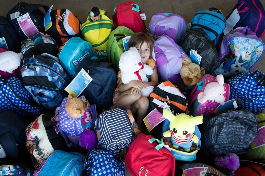 A young girl hugs a teddy smiling, lying down on a sea of colourful backpacks.