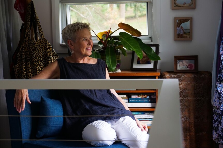 Senior-aged woman sits on couch with railing in foreground looking out window.