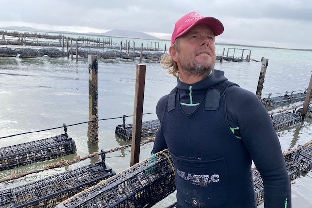 A man in a dark coloured wetsuit and red cap standing in front of an ocean holding nets