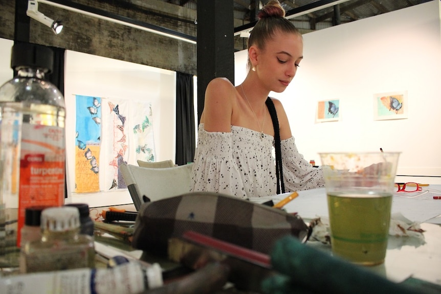 Young woman painting at desk surrounded by tubes of paint and paint brushes.