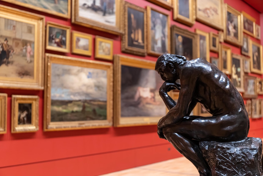 A sculpture of a man resting his head on his hands, thinking. Paintings hang on a wall in the background.