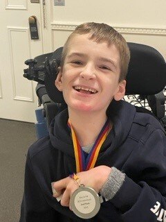A smiling boy  with a light hair, in a wheelchair holding a medal.