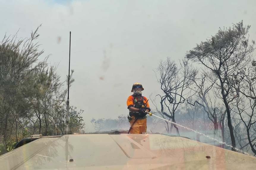 A fireman with a big white beard and sunnies holding a hose
