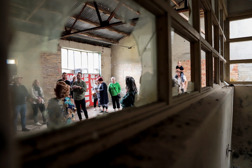 Man in centre of a group of watching people inside a dilapidated factory points to roof