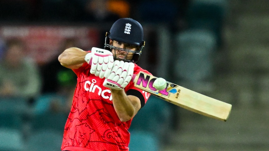 A cricket batsman, wearing a red top and a helmet, wields his bat at a delivery 