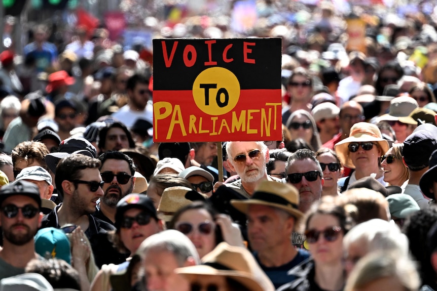 A person in a crowd holds up a sign with the Aboriginal flag and the words "Voice to Parliament". 