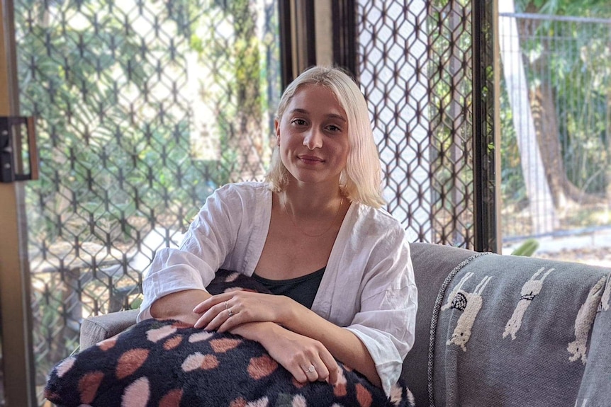 Young blonde woman sitting on couch with greenery in background.