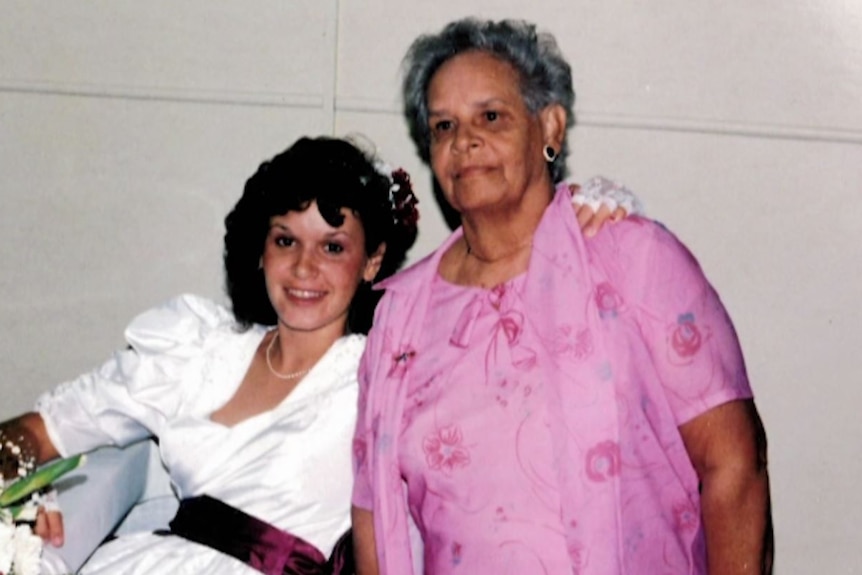 Woman in a 1980s white formal dress seated with her hand on shoulder of her mother wearing pink dress