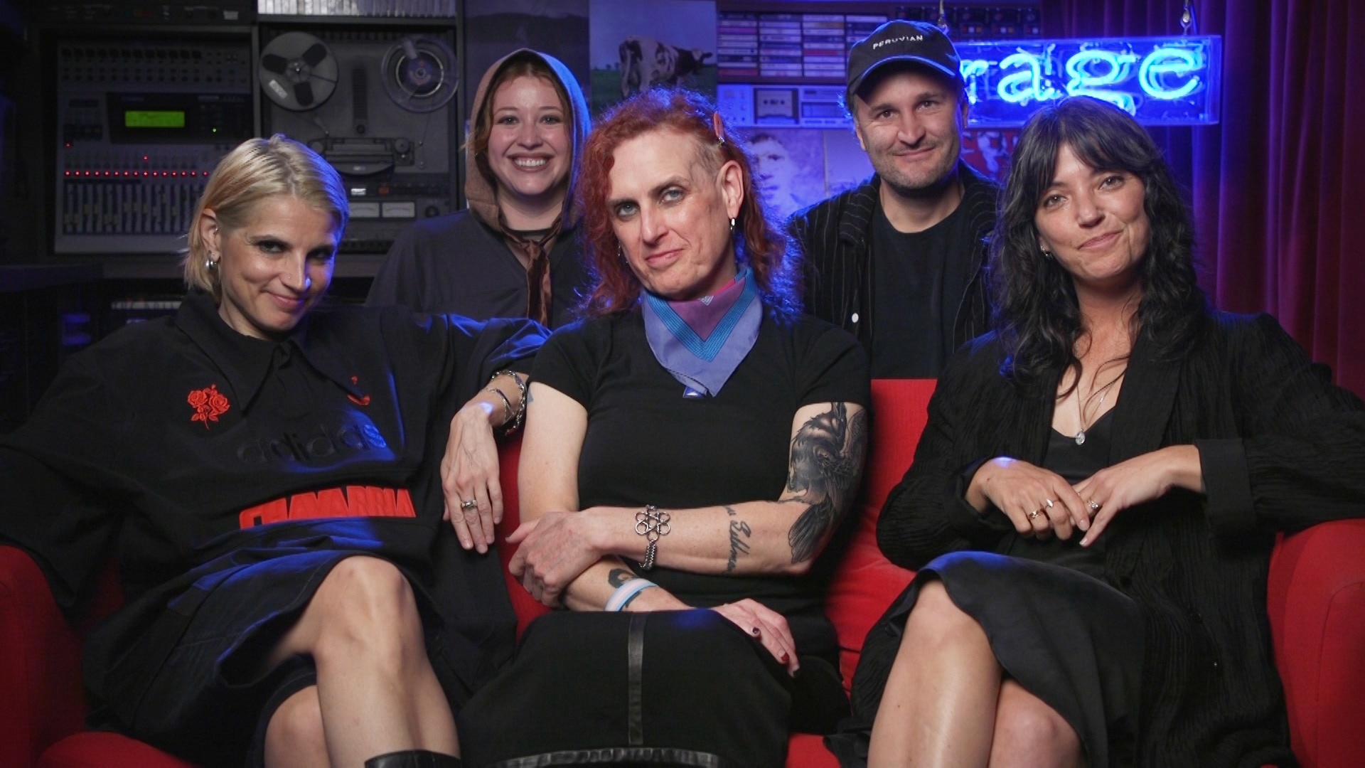 Sharon Van Etten and The Attachment Theory sit together in a dimly lit room with a red couch and neon blue sign that reads rage