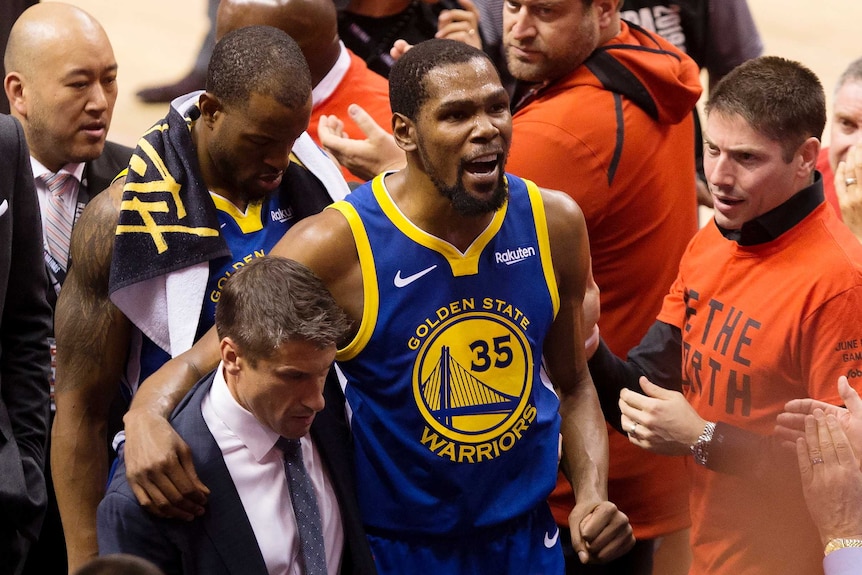 An injured NBA basketballer limps off court, assisted by officials.