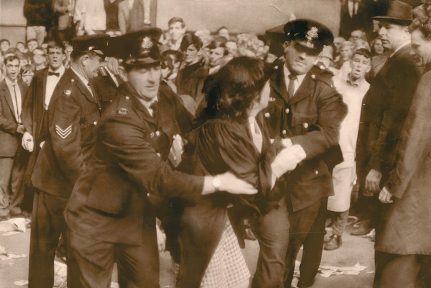 Black and white photo of police officers moving young woman away from a large crowd and she looks back.