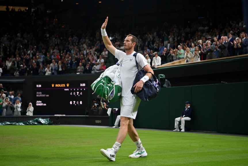 Tennis player Andy Murray carries bags on either shoulder as he raises his right hand in acknowledgement to the Wimbledon crowd.