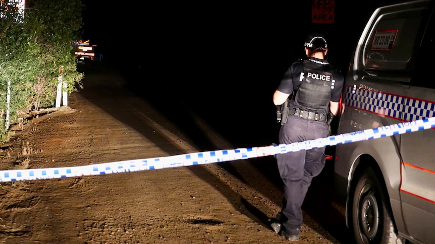 A police officer standing beside a car and cordoned off area of a secluded outback road at night