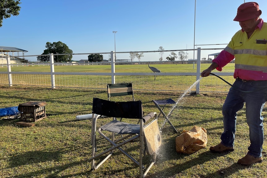 A man washes down a camping chair on the side of a football field. A brolga stands in the background.
