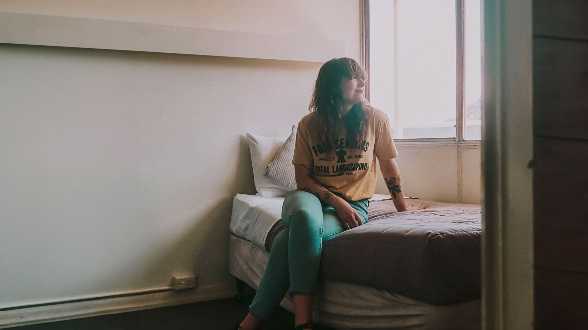 Creative Kate Berry sits on a single bed in a muted motel room and smiles, looking out the window. 