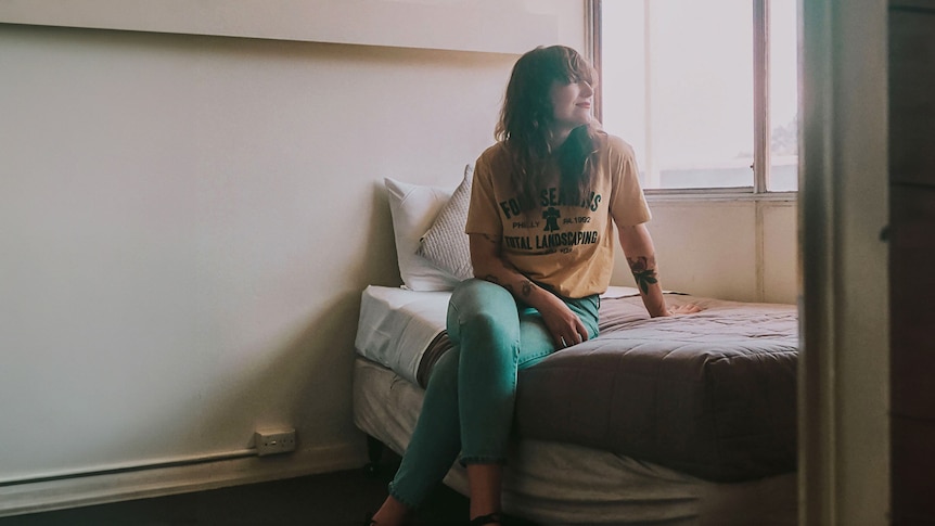 Creative Kate Berry sits on a single bed in a muted motel room and smiles, looking out the window. 