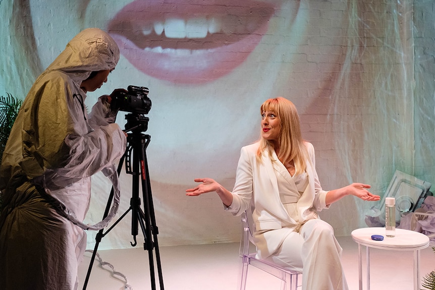 A person in white hazard suit uses camera on tripod to film Romy Bartz seated on stage in white pant suit.