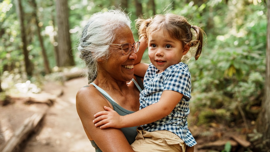 Active senior woman of Chinese Hawaiian descent with long grey hair embracing a small girl. Both grinning. In in a tall forest