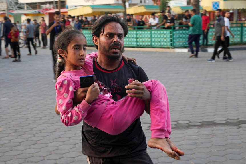 A man carries a young girl wearing pink pyjamas with a worried and urgent expression on his face