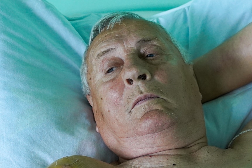 An older man lying in a hospital bed looks off to the left, resting his hand behind his head.